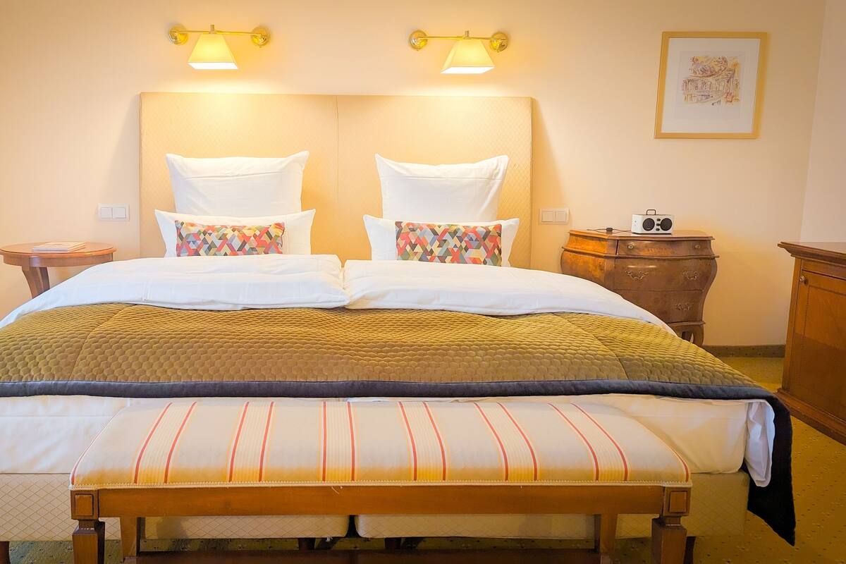 A neatly made bed with a patterned bedspread and pillows, a bedside table with a radio, and a framed picture on the wall.