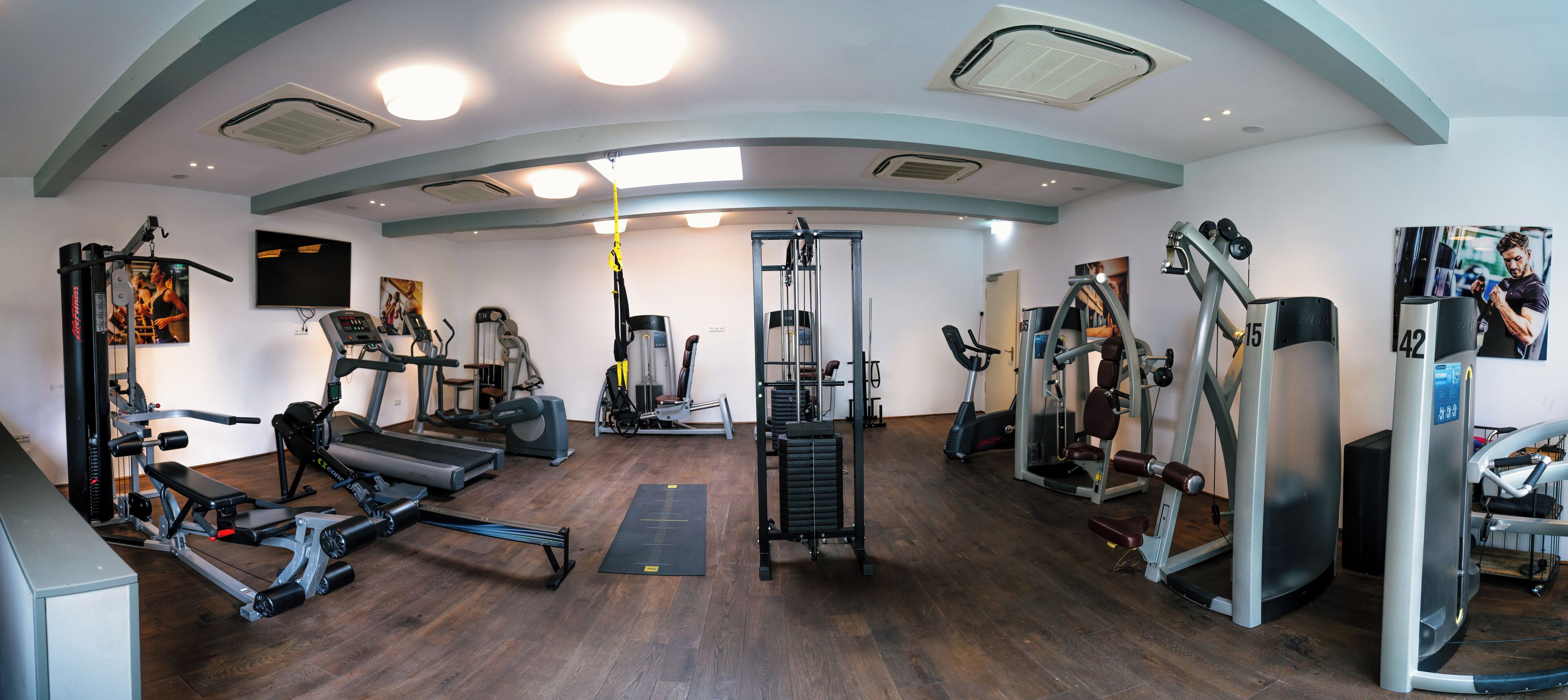 Ein Fitnessraum mit unterschiedlichen Trainingsgeräten, einem großen Spiegel und einem Holzboden.