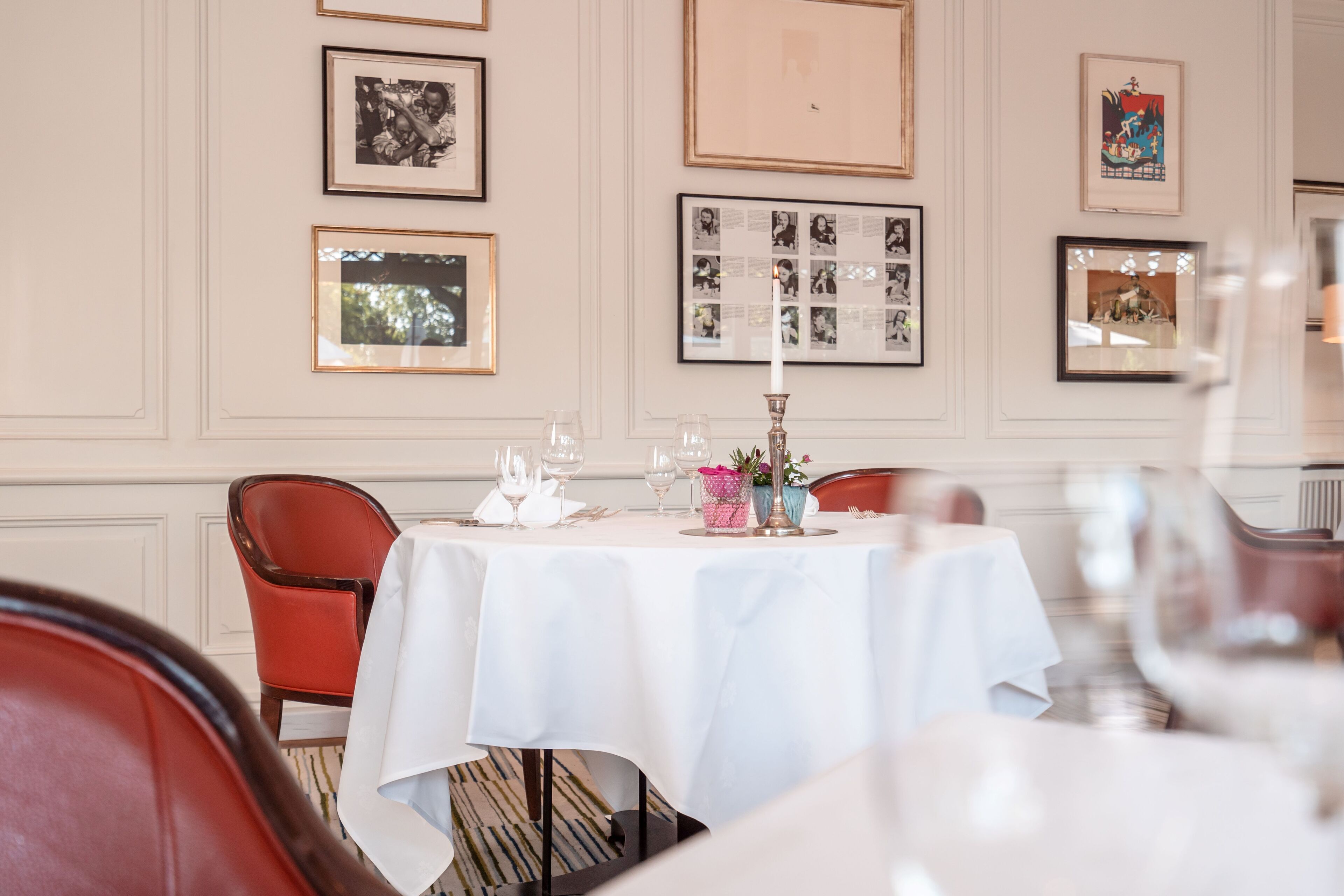 Un coin repas avec une nappe blanche, des chaises rouges, des tableaux encadrés au mur et un chandelier servant de centre de table.