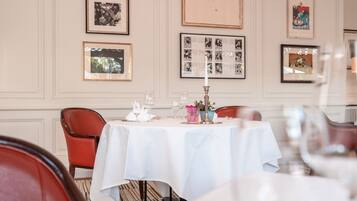 Un coin repas avec une nappe blanche, des chaises rouges, des tableaux encadrés au mur et un chandelier servant de centre de table.