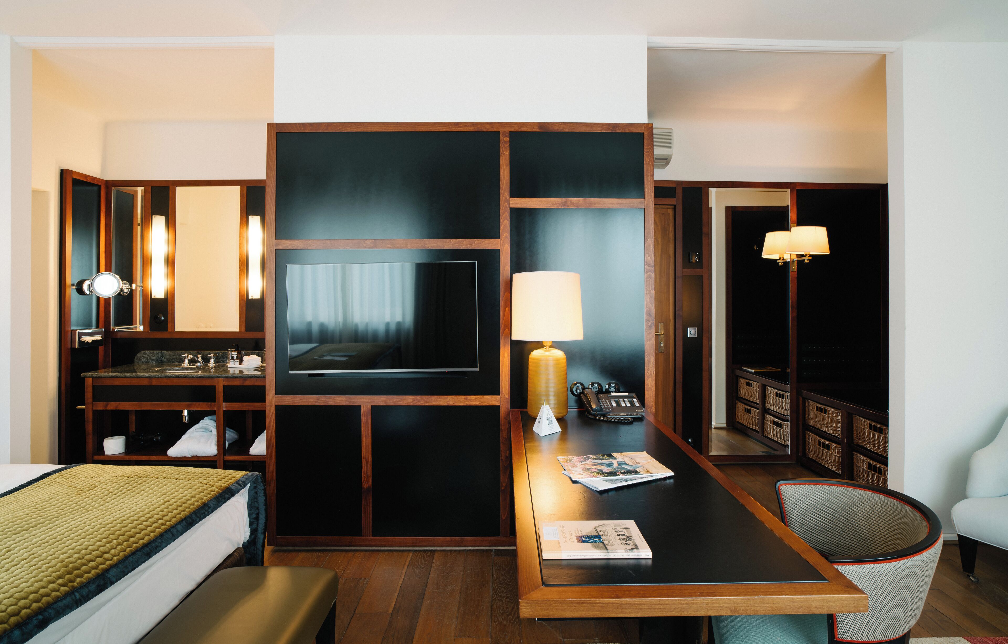 Habitación de hotel con cama, escritorio con lámpara, televisión, espejo y zona de estar con una silla.
