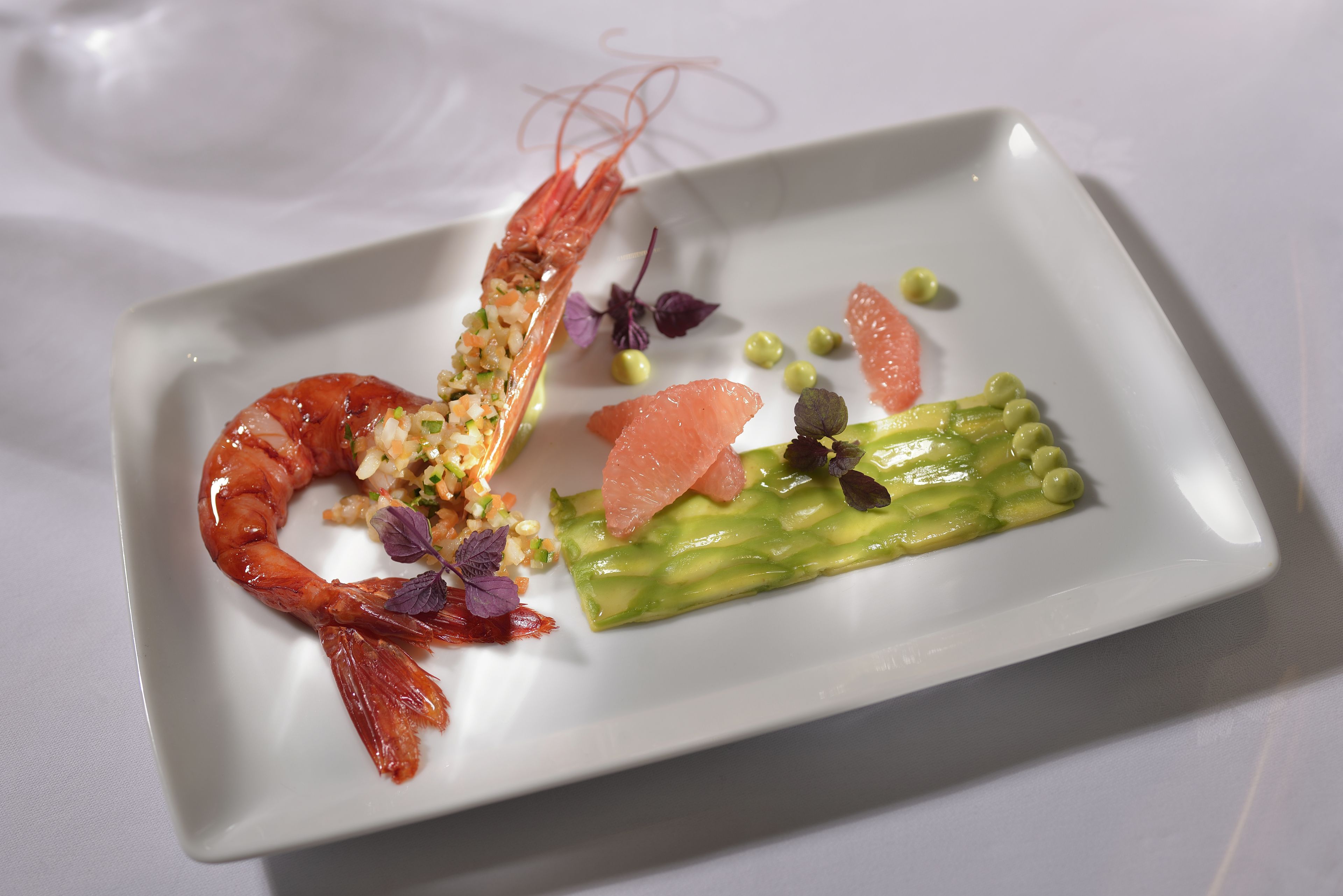 A white rectangular plate with a cooked shrimp, green asparagus, and small green peas, arranged on a white tablecloth.