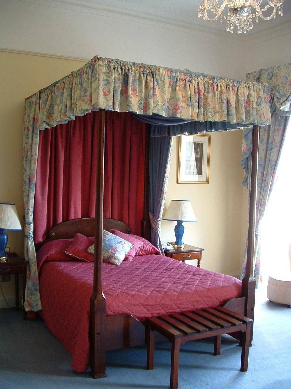 A four-poster canopy bed with red curtains, a wooden bench, a nightstand with a lamp, and a framed picture on the wall.