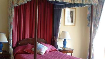 A four-poster canopy bed with red curtains, a wooden bench, a nightstand with a lamp, and a framed picture on the wall.