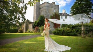 Une mariée vêtue d’une robe blanche se tient dans un champ, avec un château en arrière-plan.