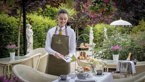 Une personne en uniforme, servant des plats à une table en plein air, avec un présentoir à gâteaux à plusieurs niveaux, le tout entouré de verdure et de statues.