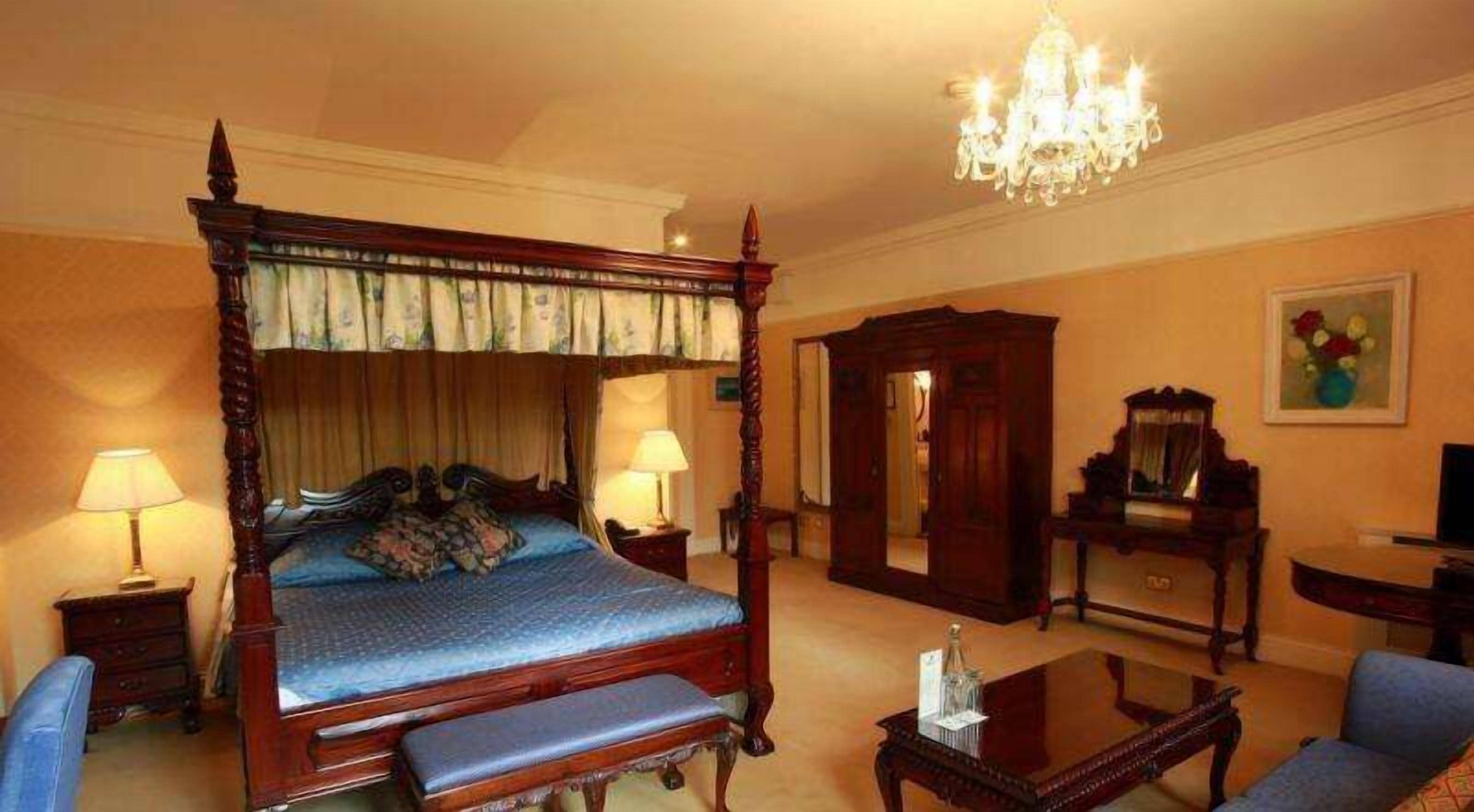 A four-poster bed with a canopy, a chandelier, a desk, and a chair in a hotel room.