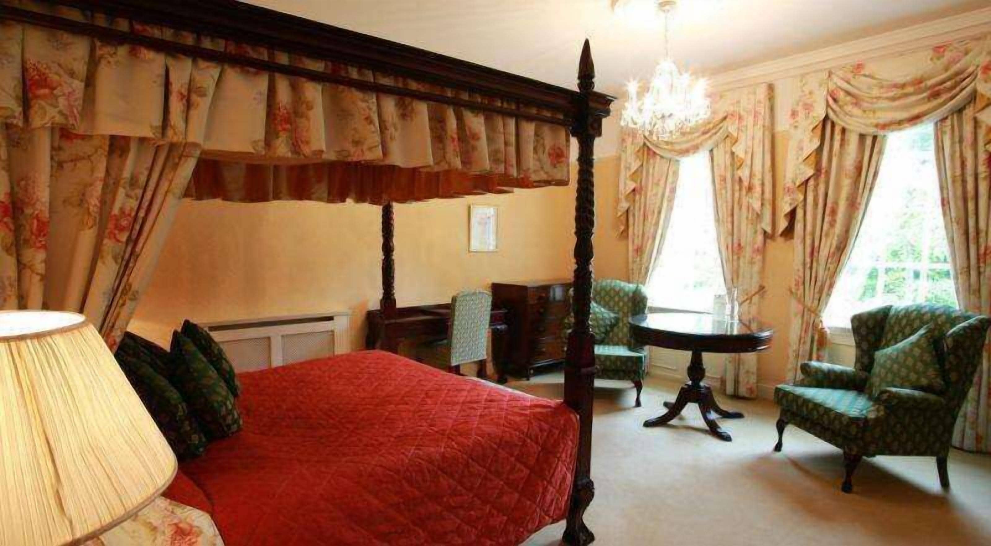 A four-poster bed with a red bedspread, a chandelier, floral curtains, and two armchairs.