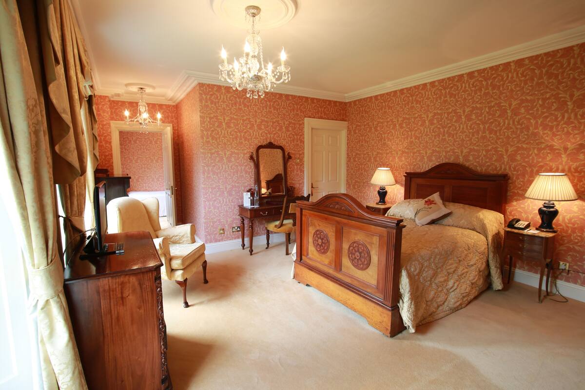 Quarto com cama grande, cômoda de madeira, cadeira, escrivaninha, espelho e lustre.