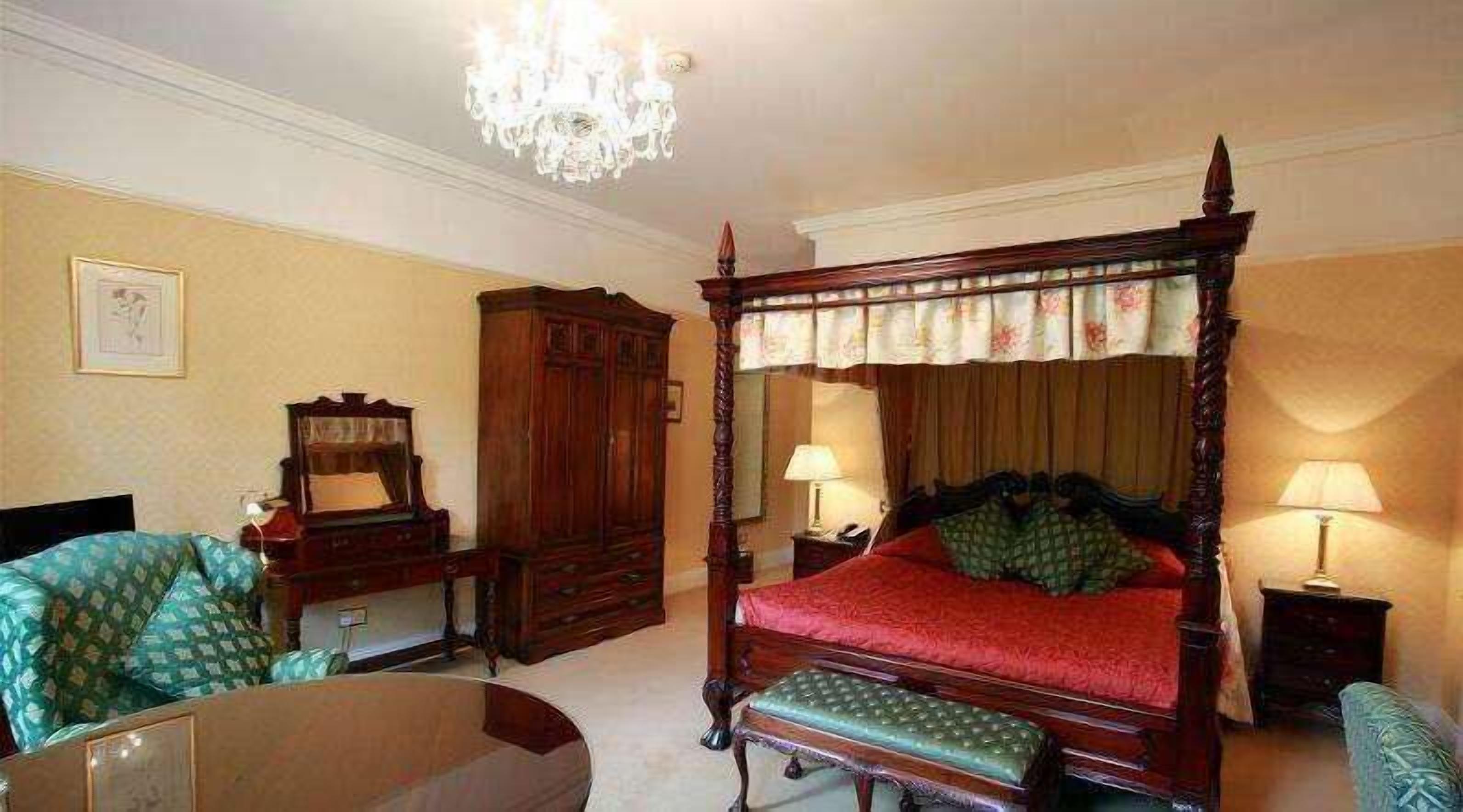 A four-poster bed with a red bedspread, a wooden nightstand with a lamp, a green upholstered armchair, and a wooden cabinet.