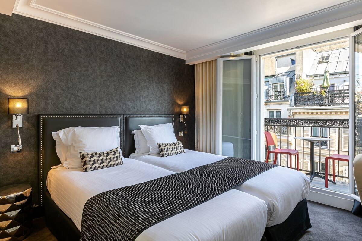 Een hotelkamer met twee bedden, een donkere wand en een balkon met rode stoelen en een tafel.