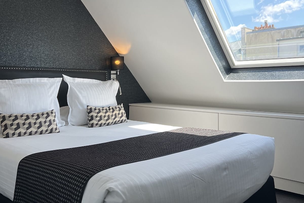 Een moderne slaapkamer met een schuine kap, dakraam, een bed met witte lakens, patroon kussens en een zwart accentkussen.