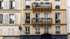Un edificio de varios pisos con un estilo arquitectónico clásico, con balcones adornados con plantas, un escaparate visible con señalización y una motocicleta estacionada en la acera.