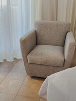 Un fauteuil beige dans une pièce dont le sol est carrelé et où sont accrochées des rideaux blancs.
