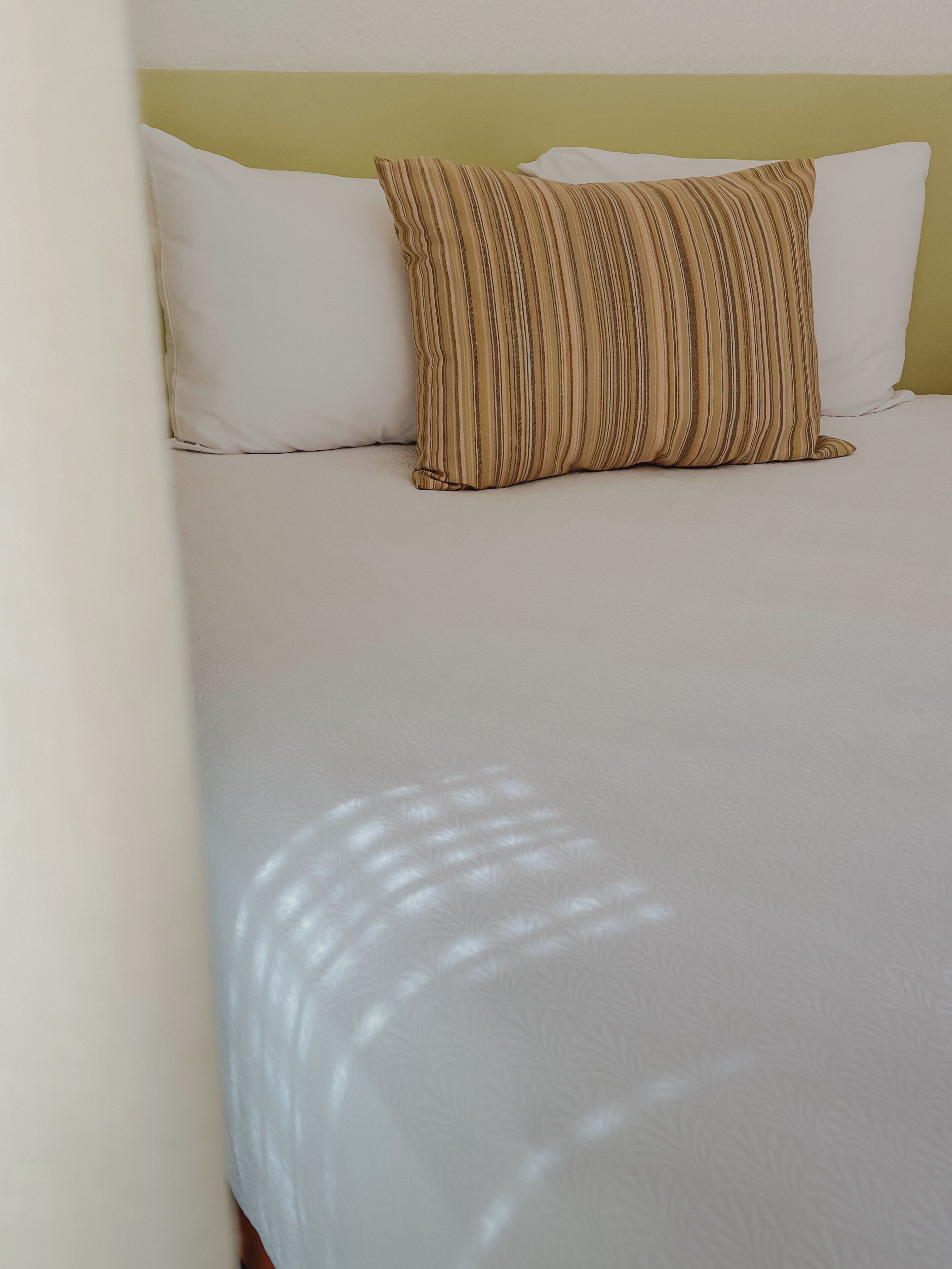 Una cama con ropa de cama blanca y una almohada a rayas, una pared lisa y una pared de acento amarilla.