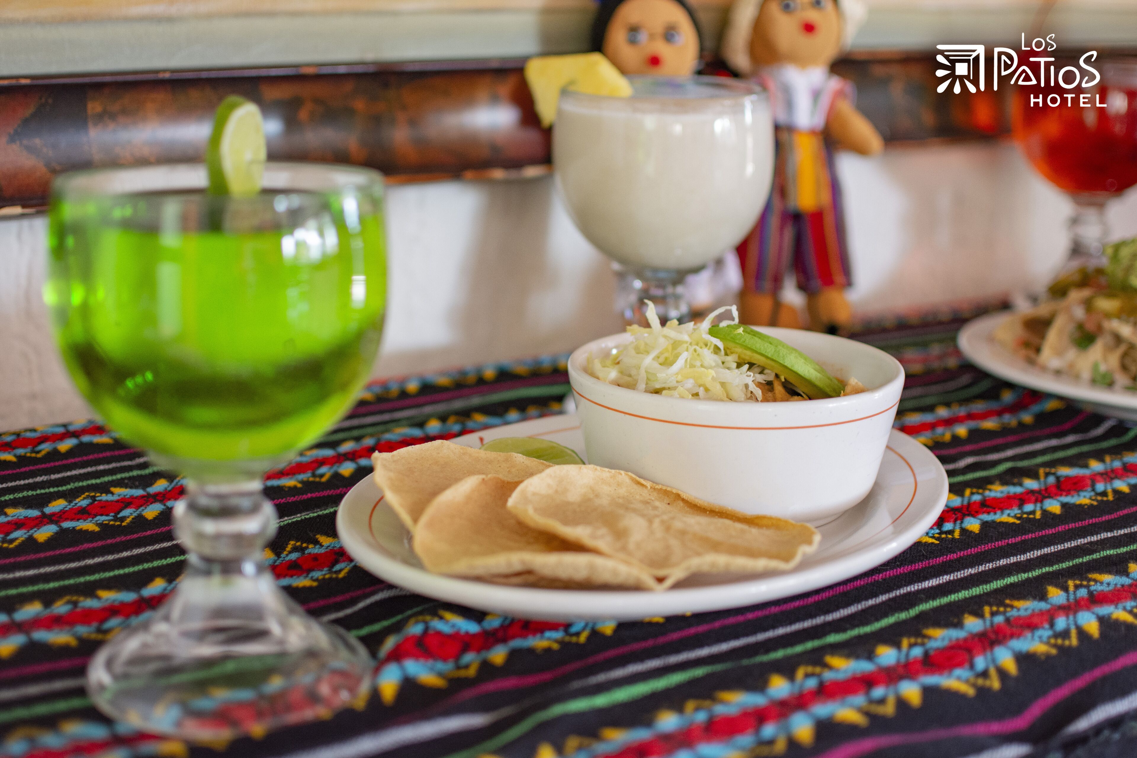 A table with a colorful tablecloth, a bowl of food, a glass of green drink, and a doll in the background.