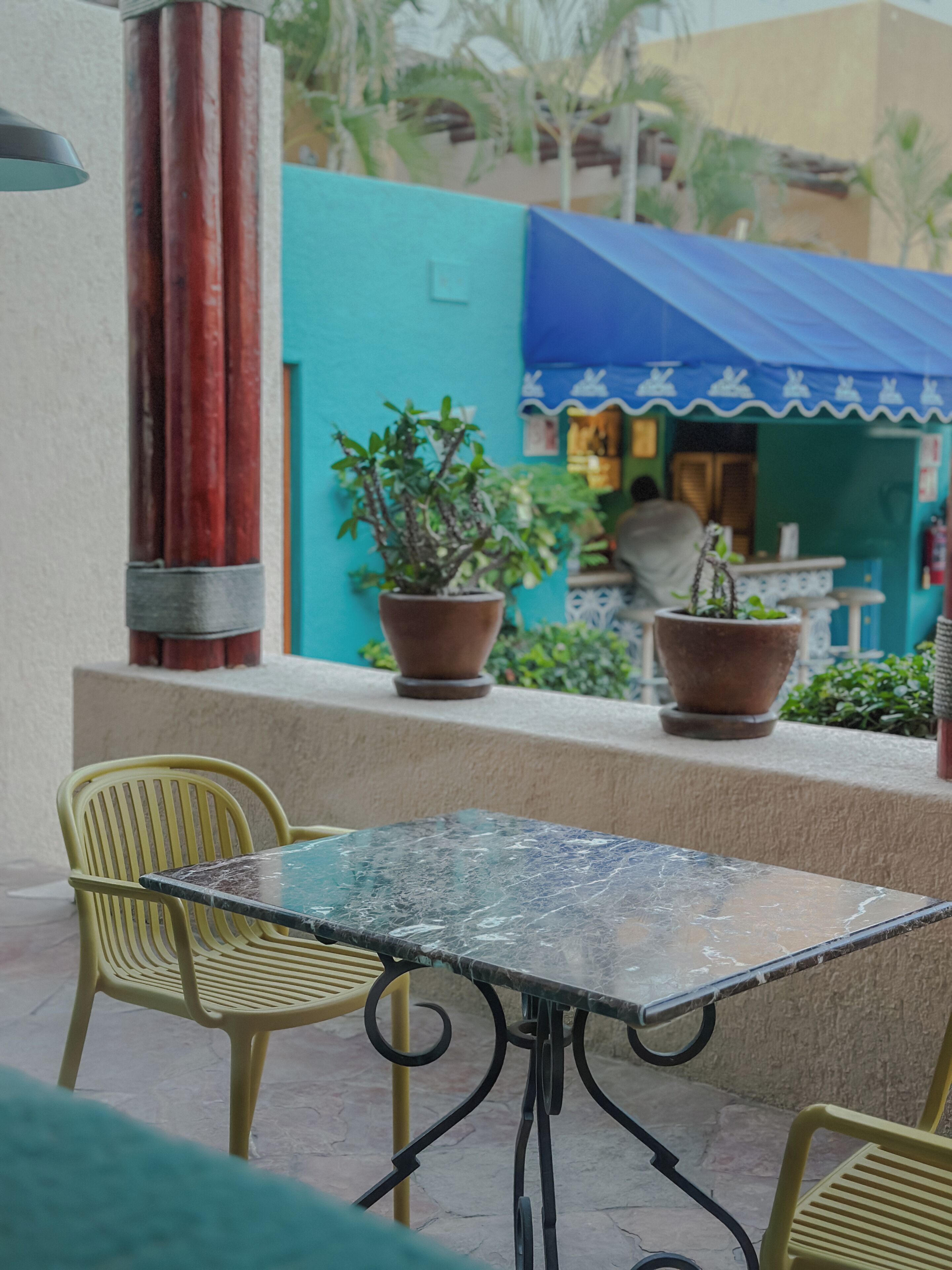 Un espacio al aire libre con mesa y sillas, plantas en macetas y un edificio con toldo azul al fondo.