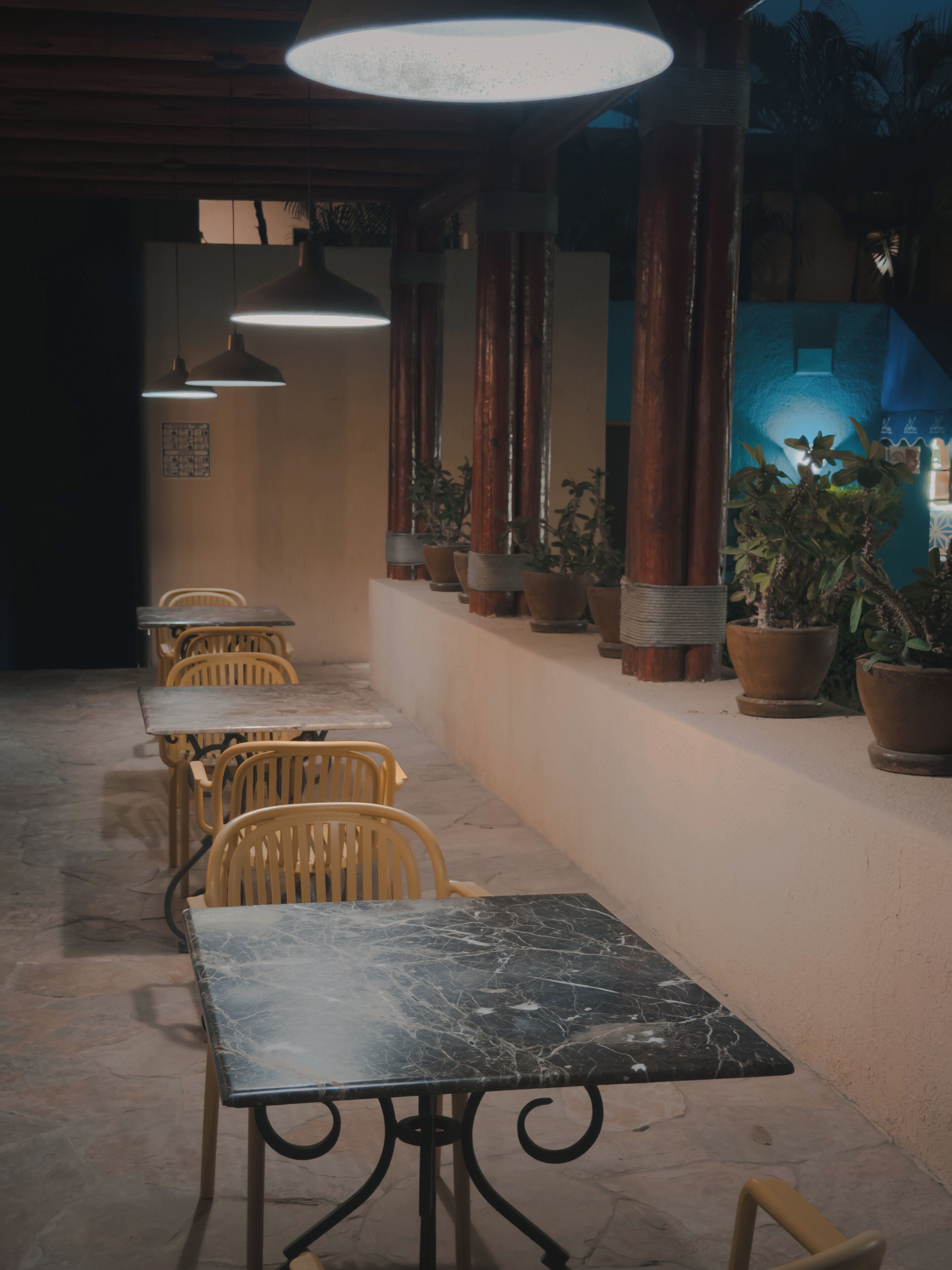Un restaurante con mesas y sillas de mármol, plantas en macetas y lámparas colgantes.