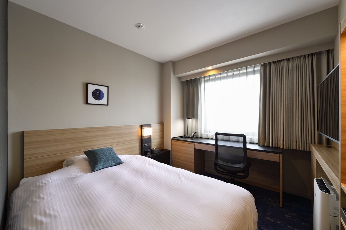 Quarto de hotel com cama, escrivaninha, cadeira e janela com cortinas.