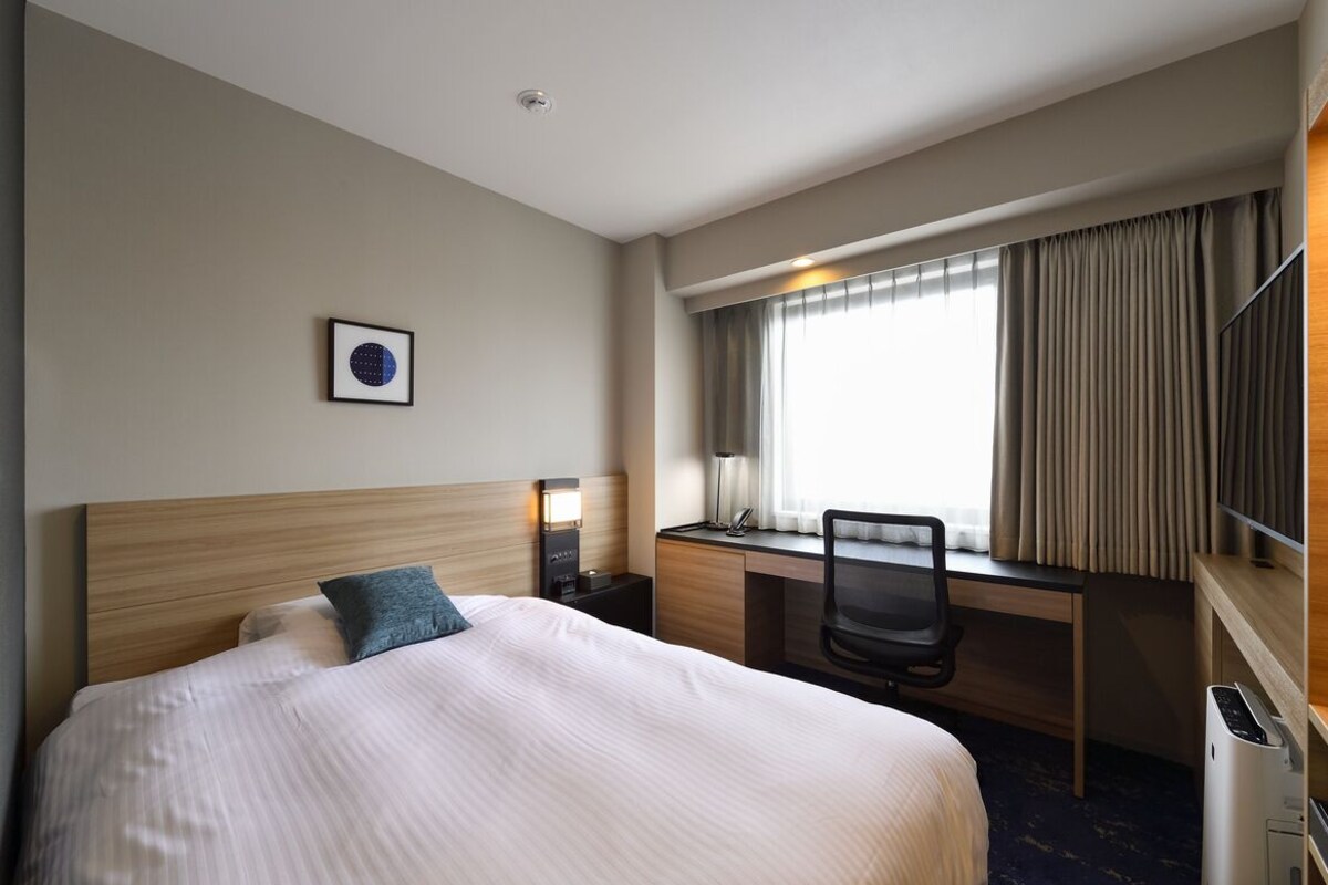 A hotel room with a bed, desk, chair, and a window with curtains.
