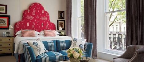 A bedroom with a large bed, a sofa, a coffee table, and a chair. There is a window with curtains, a lamp, and framed pictures on the wall.