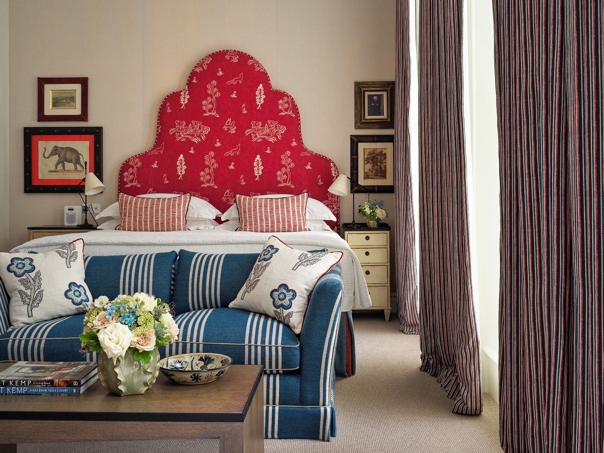 A neatly arranged bedroom with a striped sofa, a red headboard, and a bedside table with books and a vase.