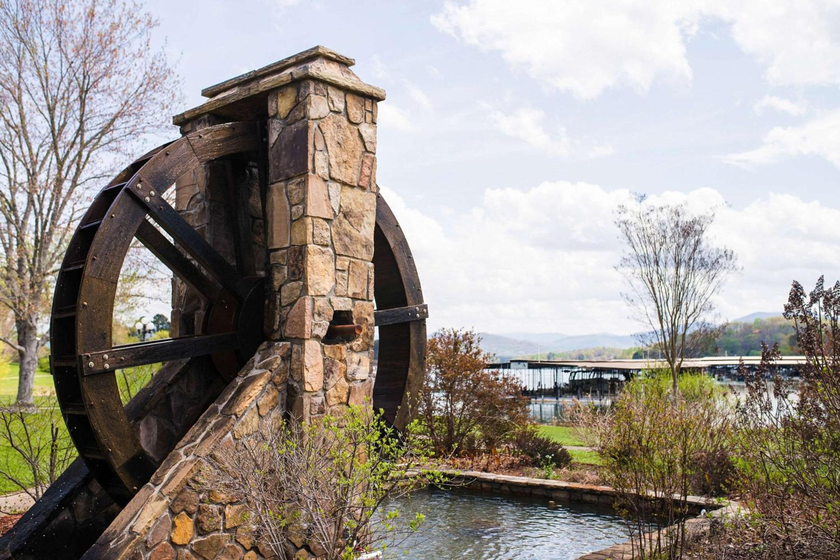 A large water wheel structure with stone architecture, set in a landscaped area with a body of water and a bridge in the background.