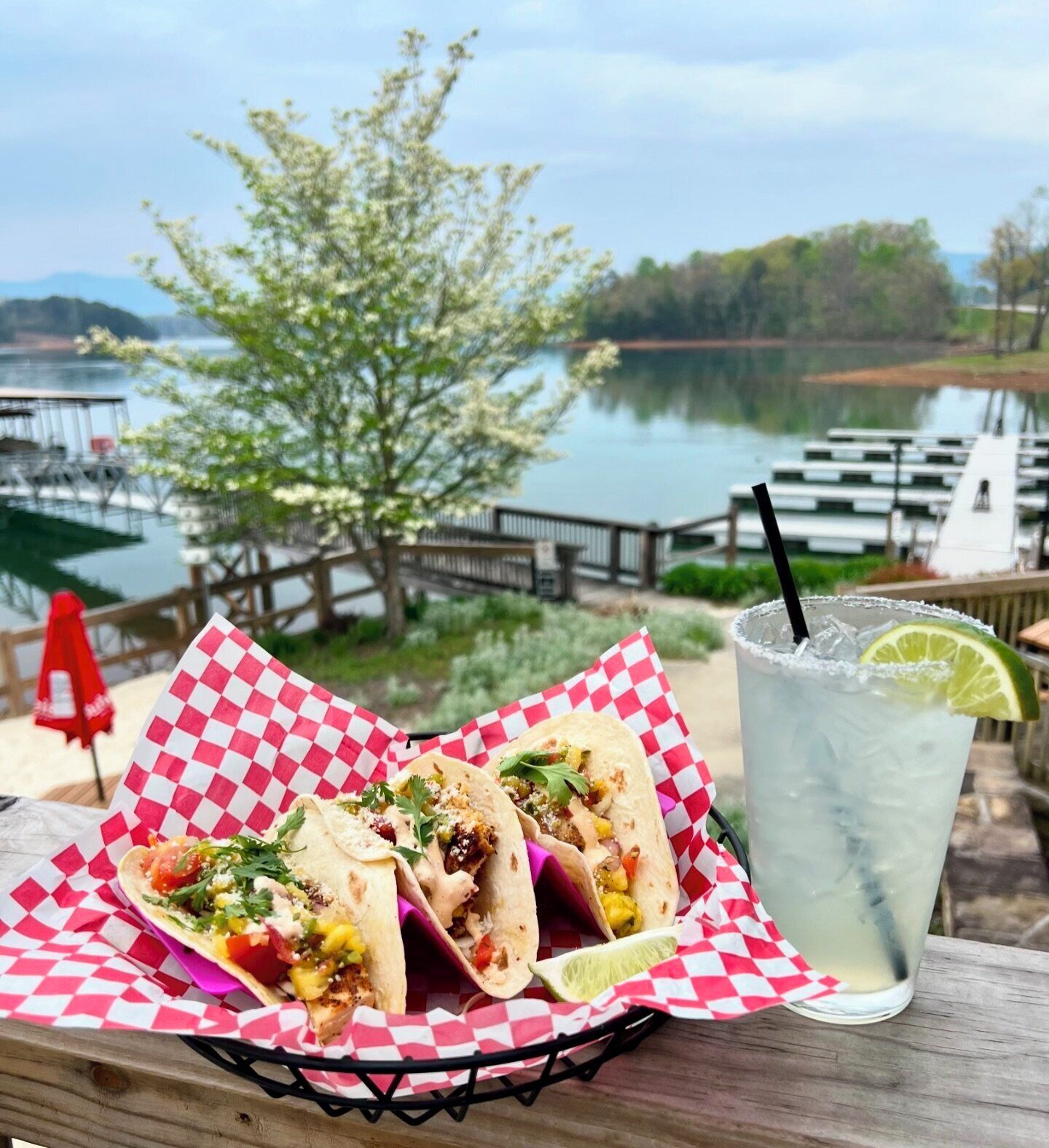 A wooden table with a basket of tacos and a glass of iced drink with a lime wedge.