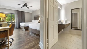 Habitación de hotel con escritorio, dos camas, ventilador de techo, baño con espejo y vistas a zonas verdes.