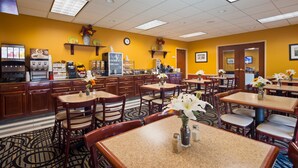 A hotel dining area with multiple tables and chairs, a kitchen area with coffee machines, and decorative vases with flowers.