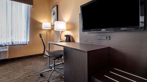 A hotel room with a desk, chair, TV, lamps, and a window with curtains.
