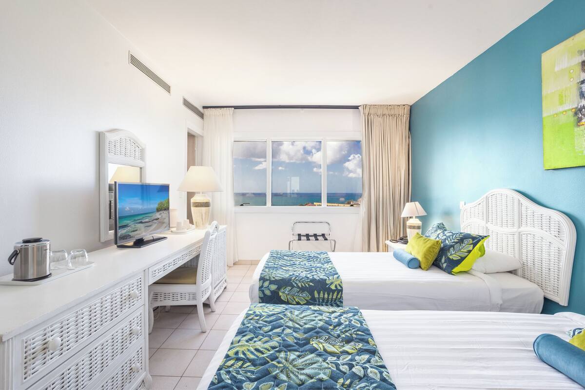 A hotel room with two beds, a desk with a TV, a window with a view, and a white headboard.
