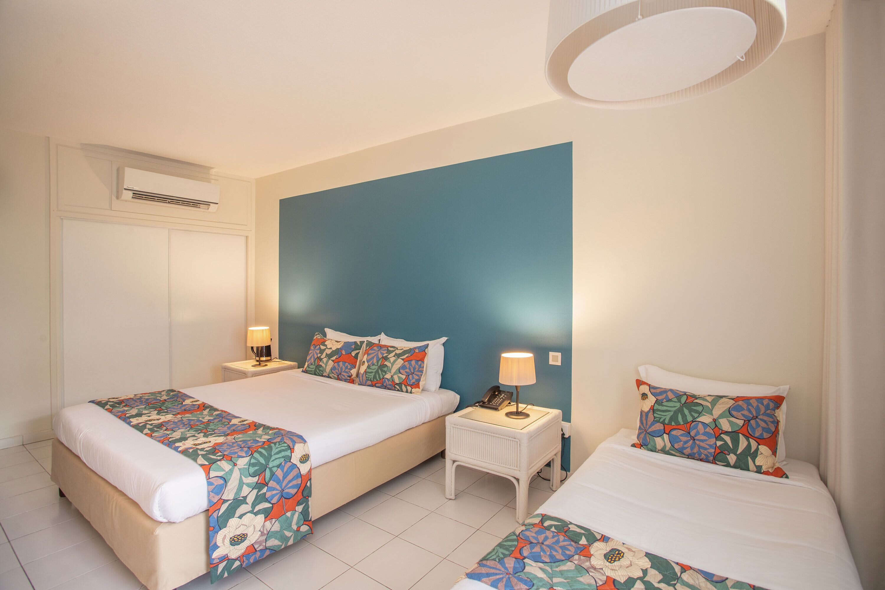 Une chambre d’hôtel avec deux lits, une table de chevet avec une lampe, un appareil de climatisation et un mur d’accent bleu.
