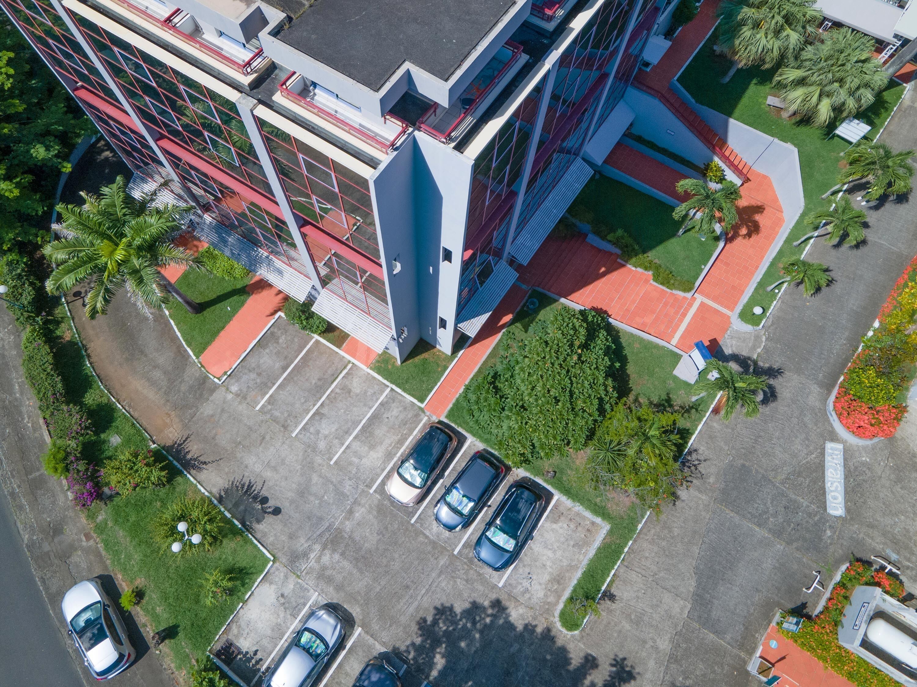 An aerial view of a modern building with a parking lot, green spaces, and a few parked cars.