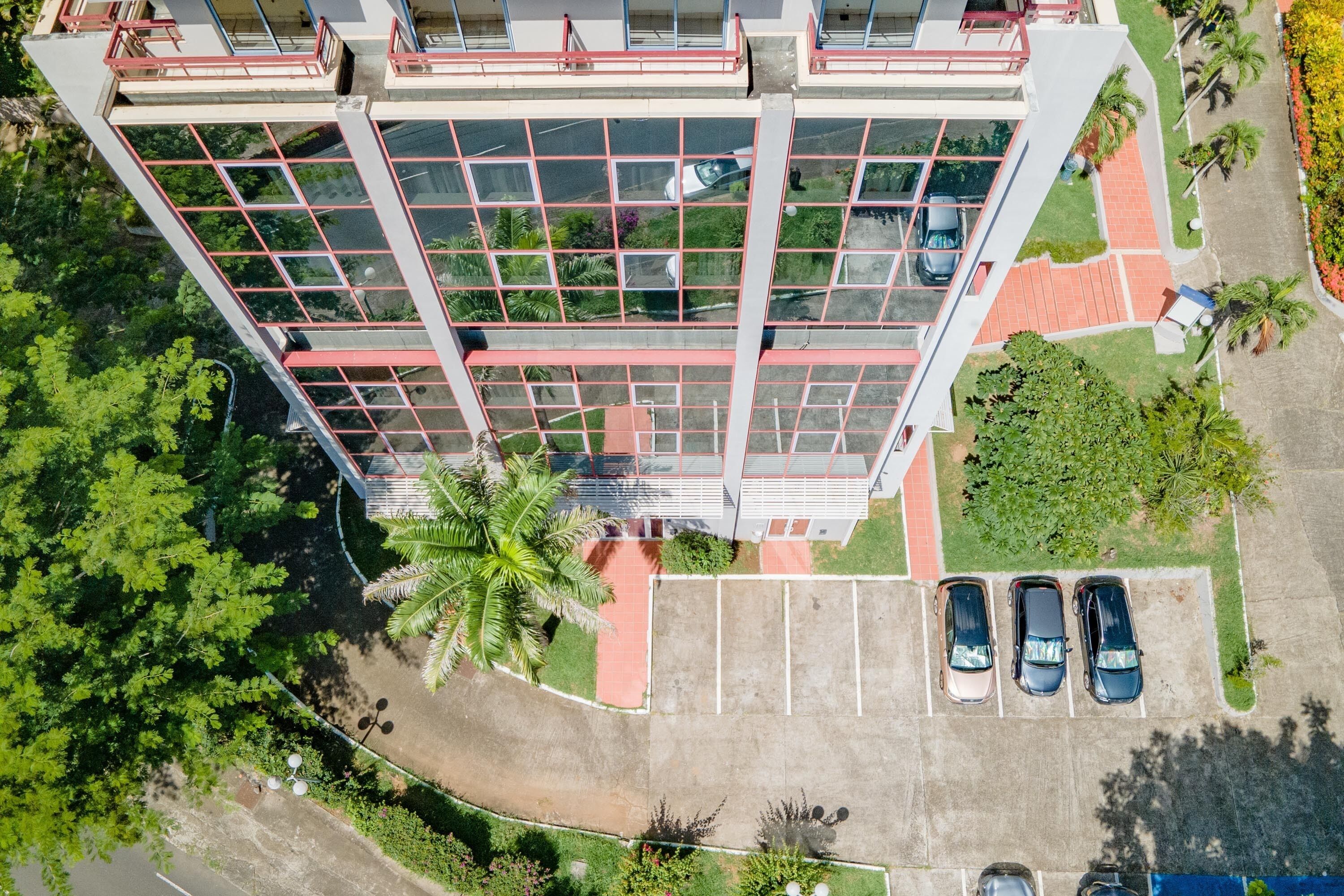 A modern building with a glass facade, a parking area with three cars, and surrounding greenery.