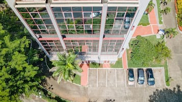 A modern building with a glass facade, a parking area with three cars, and surrounding greenery.