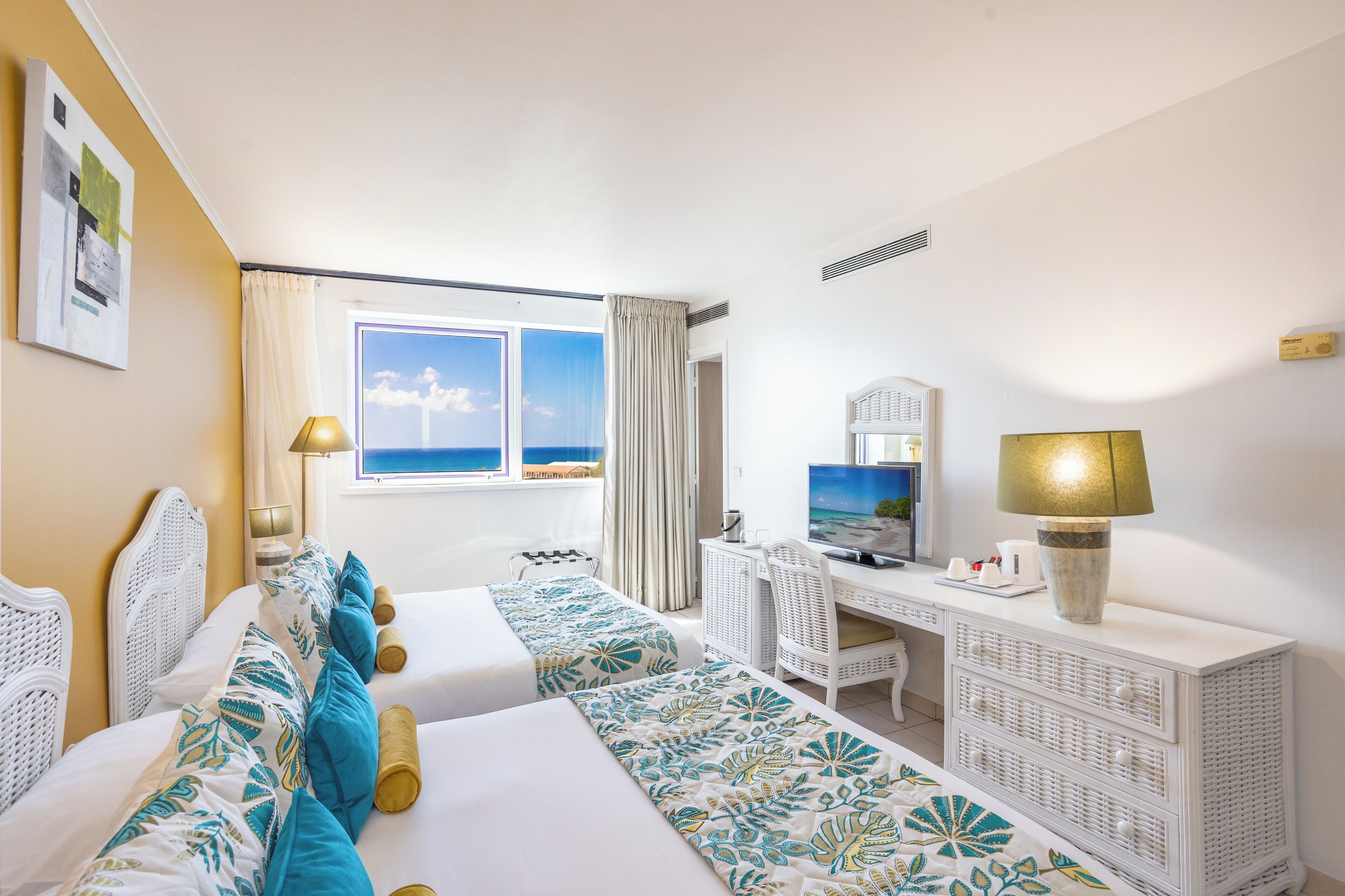 A hotel room with two beds, a desk, a chair, a television, and a window with a view of the sea.