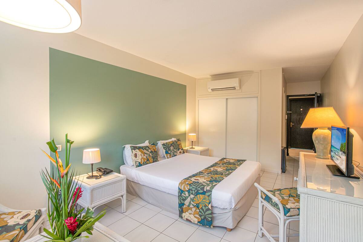 A hotel room with a bed, bedside tables, a desk, a chair, a lamp, a plant, and an air conditioning unit.