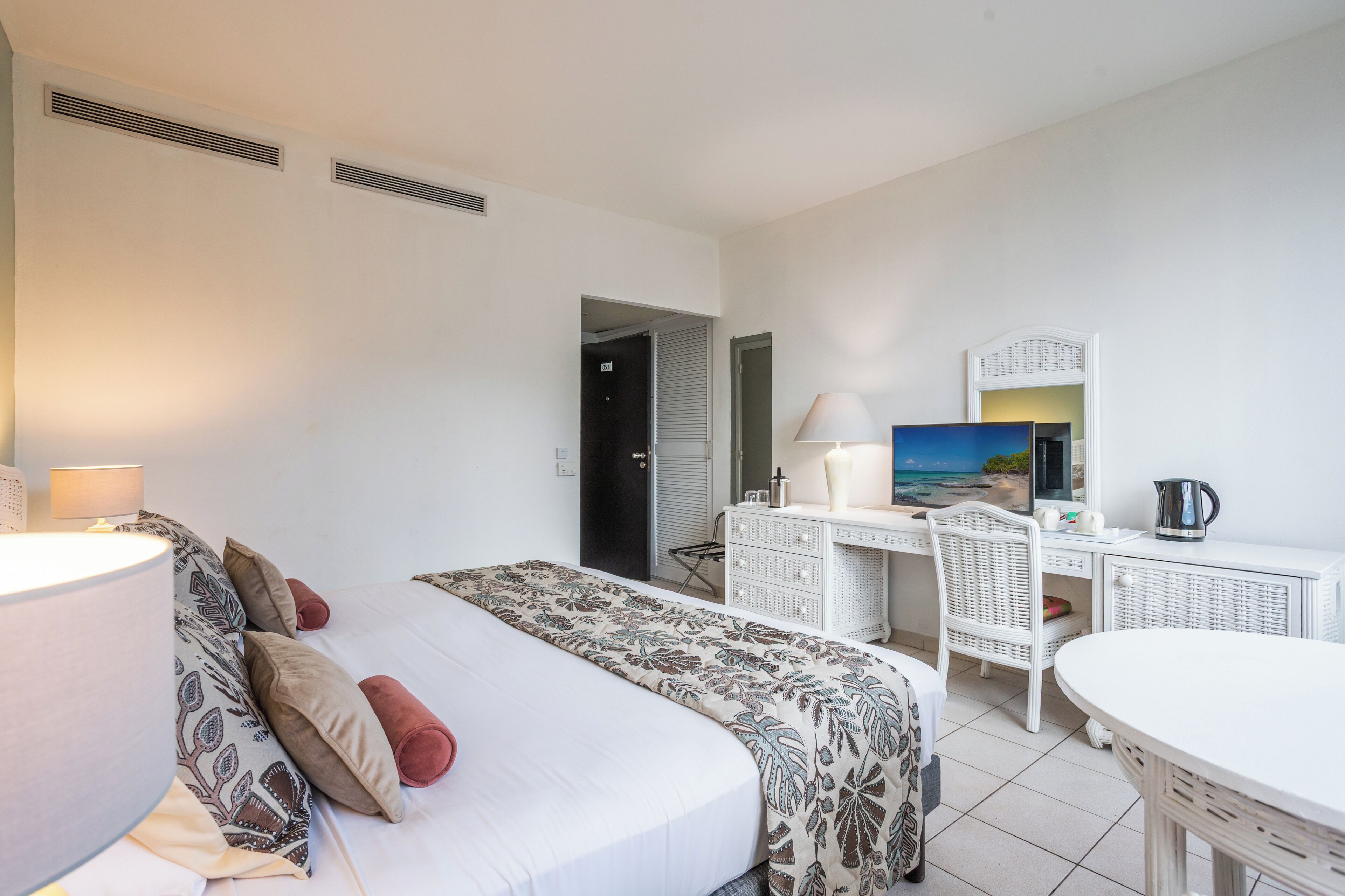 A hotel room with a bed, a desk, a chair, a lamp, a painting, and a kettle.