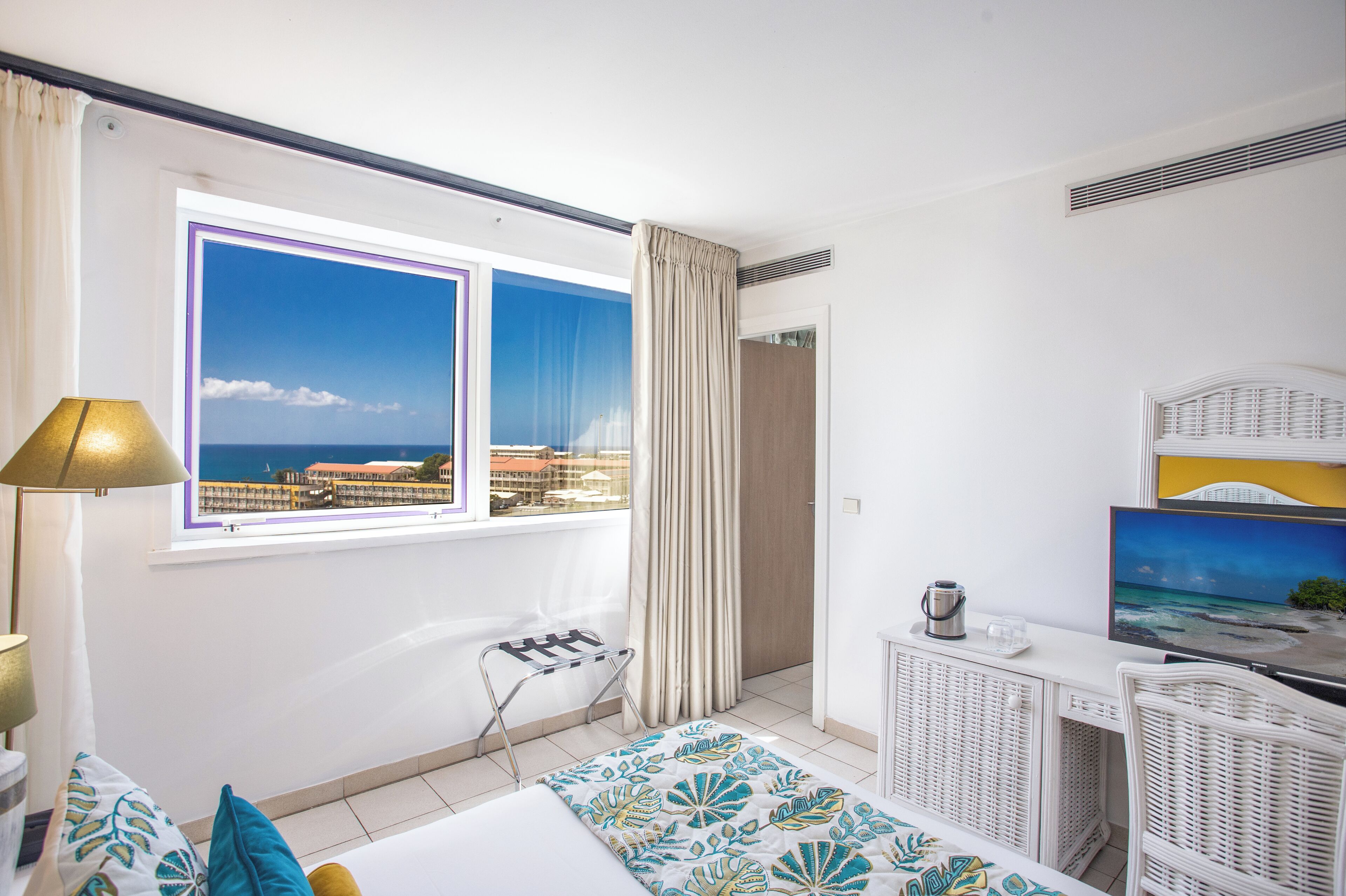 A hotel room with a bed, a TV, a window with a view of the sea, a lamp, and a small table with a kettle.