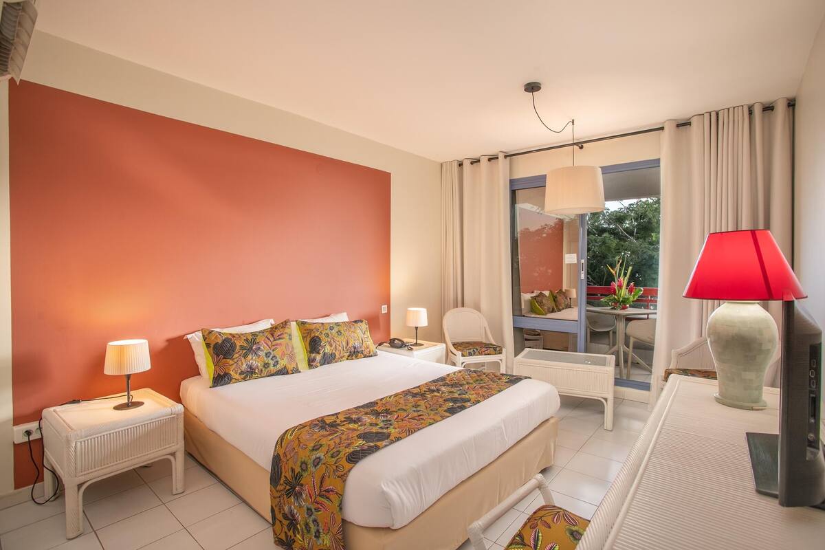 A hotel room with a bed, a nightstand, a desk with a lamp, a chair, and a view of the outdoors.