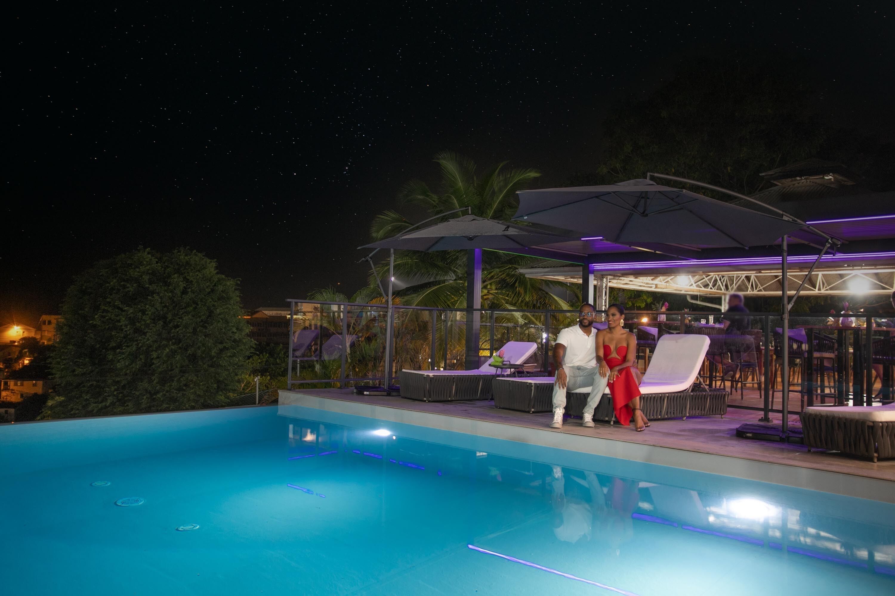 A swimming pool with lounge chairs and umbrellas, surrounded by palm trees and a night sky.