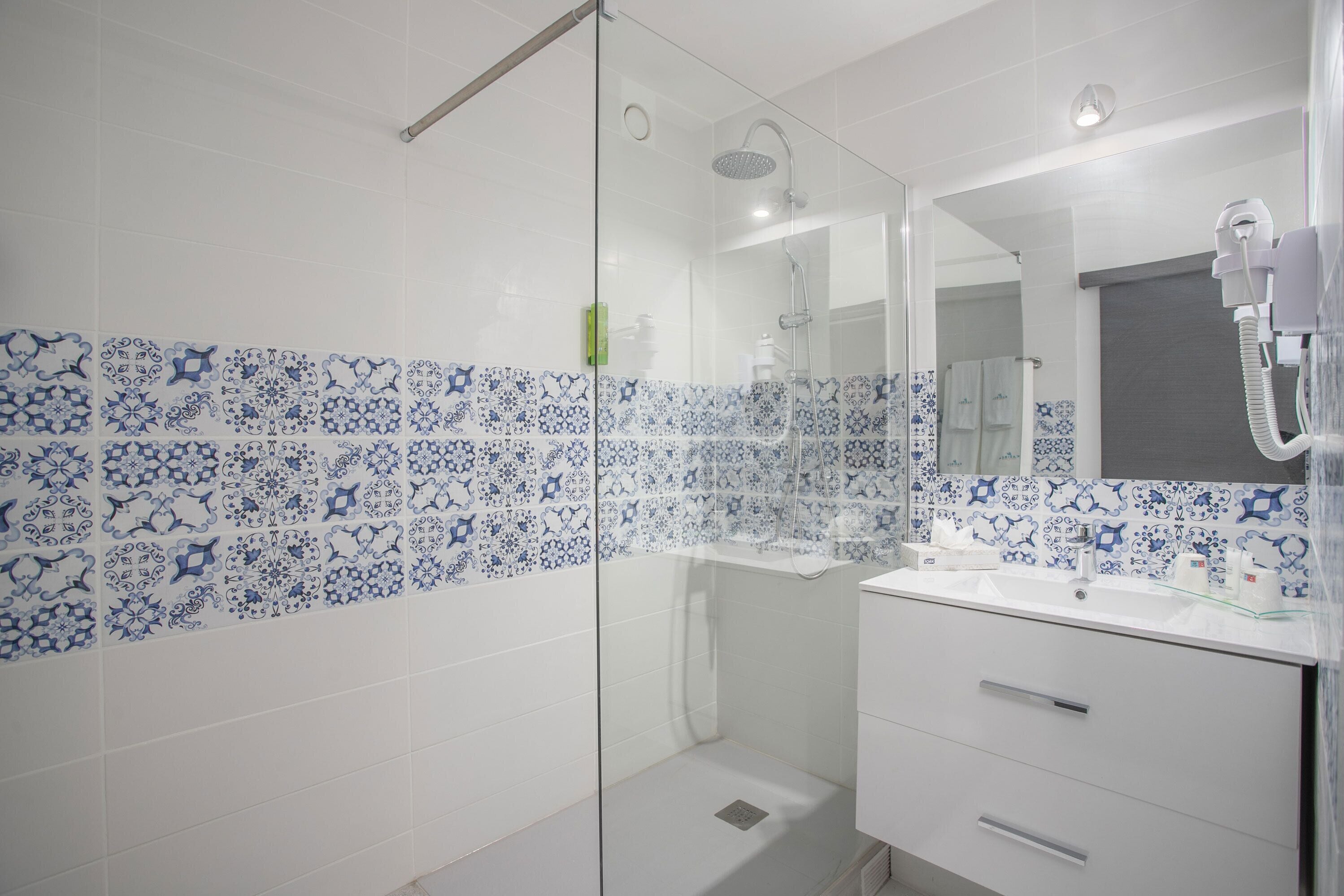 Un baño con cabina de ducha de vidrio, azulejos blancos con motivos florales azules, un lavabo blanco y un espejo con secador de pelo.