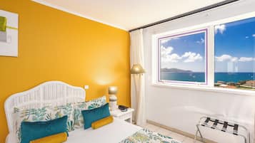 Quarto com cama, mesa de cabeceira, luminária, cadeira e janela com vista para o mar.