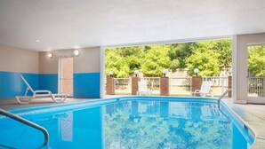 An indoor swimming pool with clear blue water, white lounge chairs, and a view of a brick wall and greenery outside.