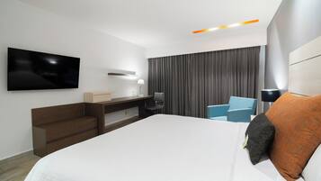 Quarto de hotel moderno com uma cama grande, televisão de tela plana na parede, uma escrivaninha com cadeira e um espaço de estar com duas poltronas.