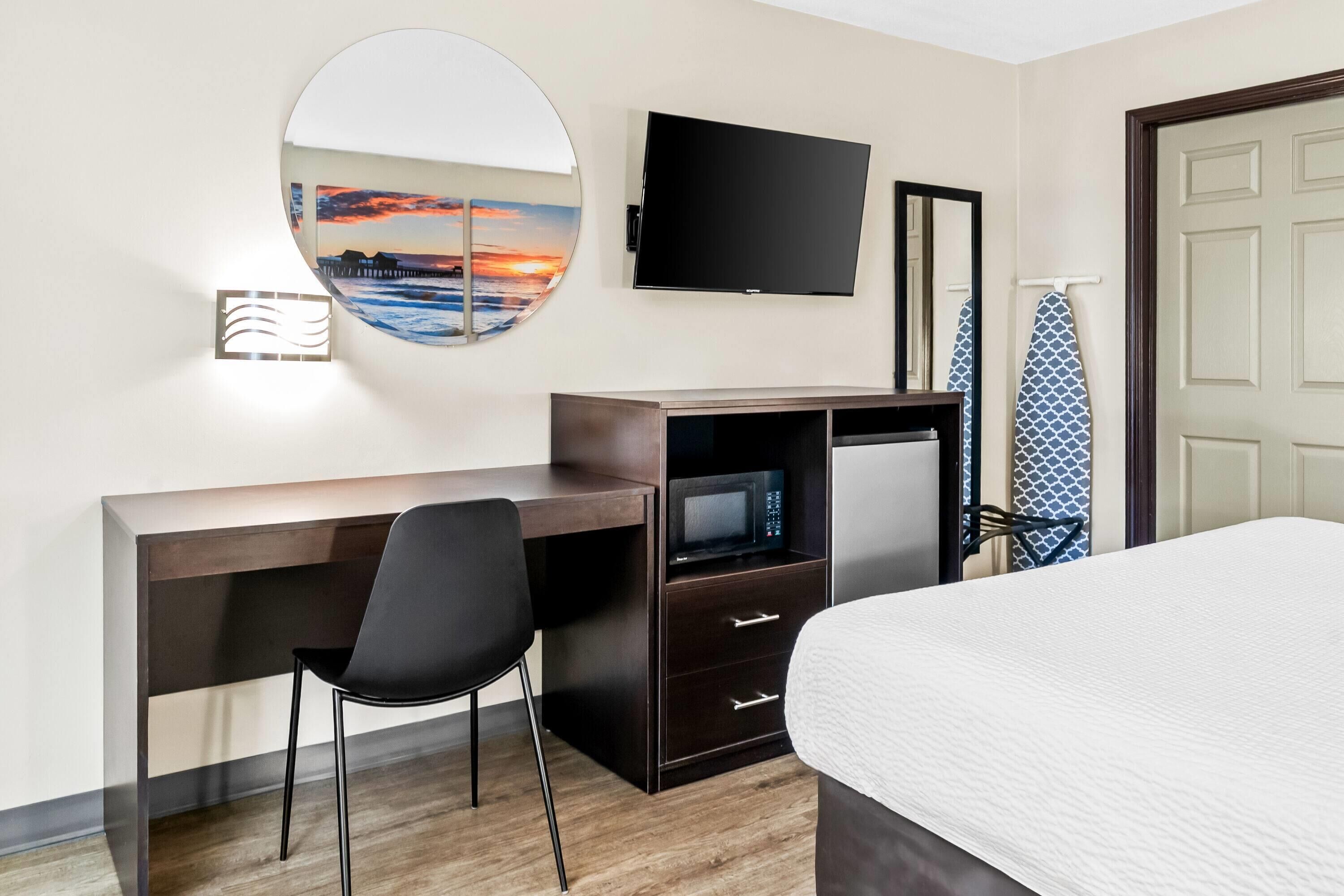 Una habitación de hotel con una cama, un escritorio con una silla, un televisor de pantalla plana, un microondas, un espejo y una puerta con un perchero para toallas.
