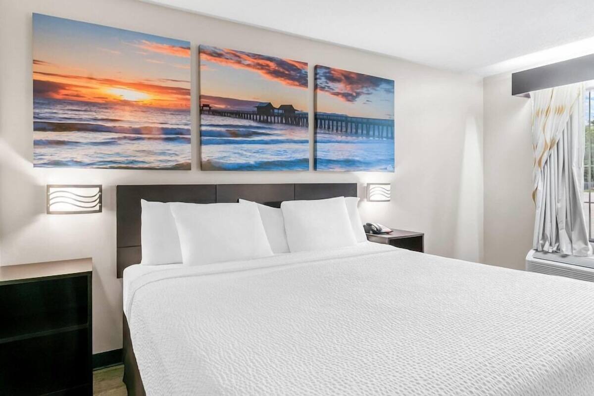 A hotel room with a large bed, a nightstand, a wall-mounted lamp, and a view of the ocean with a pier.