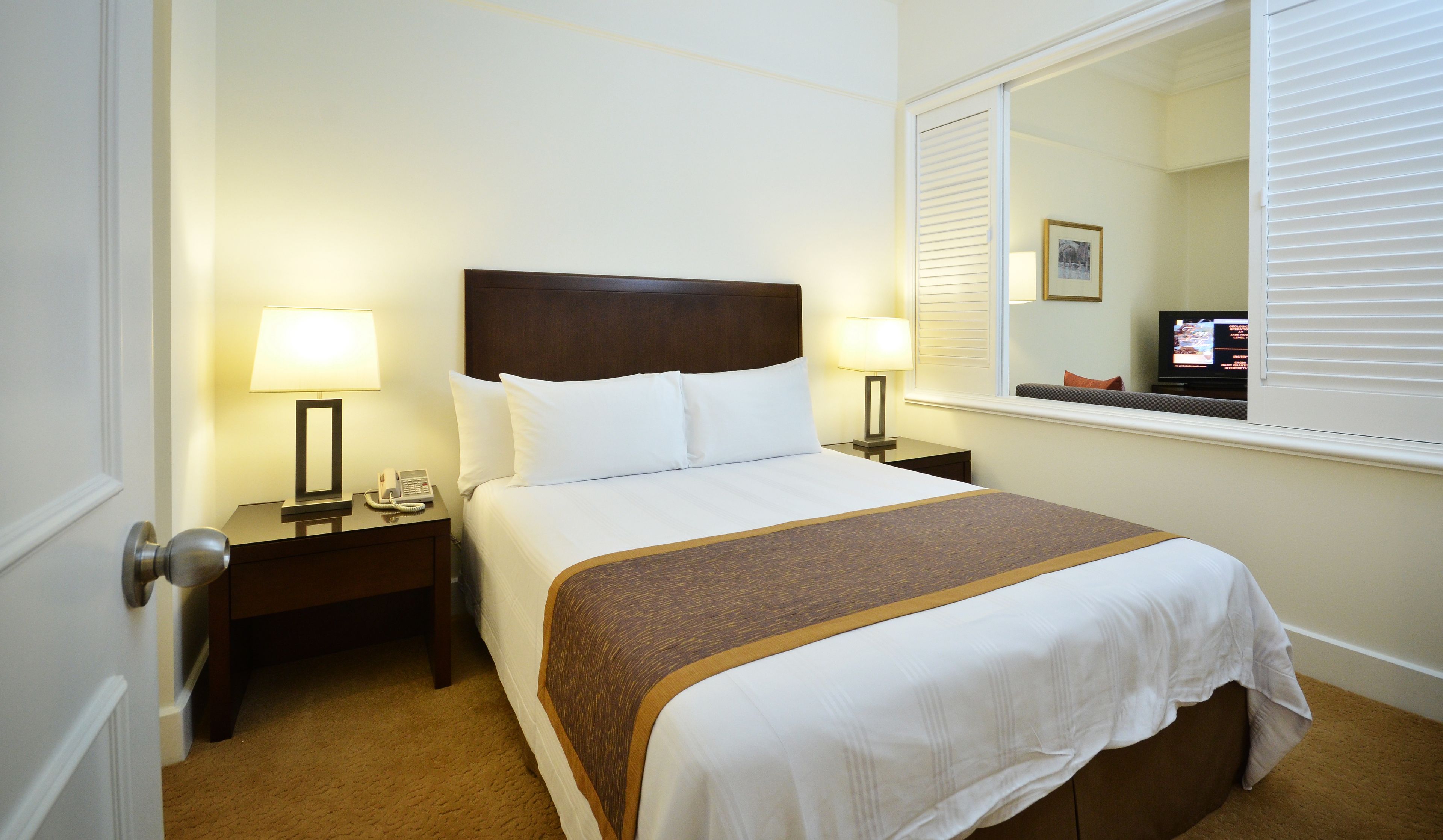 A hotel room with a bed, two bedside tables with lamps, a nightstand, a window with blinds, and a television.