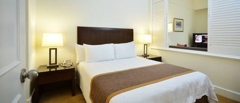 A hotel room with a bed, two bedside tables with lamps, a nightstand, a window with blinds, and a television.